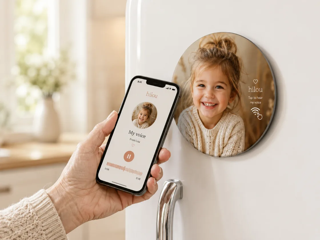 A hand holds a phone showing the hilou audio player next to a round photo magnet on the fridge, playing the child's voice message